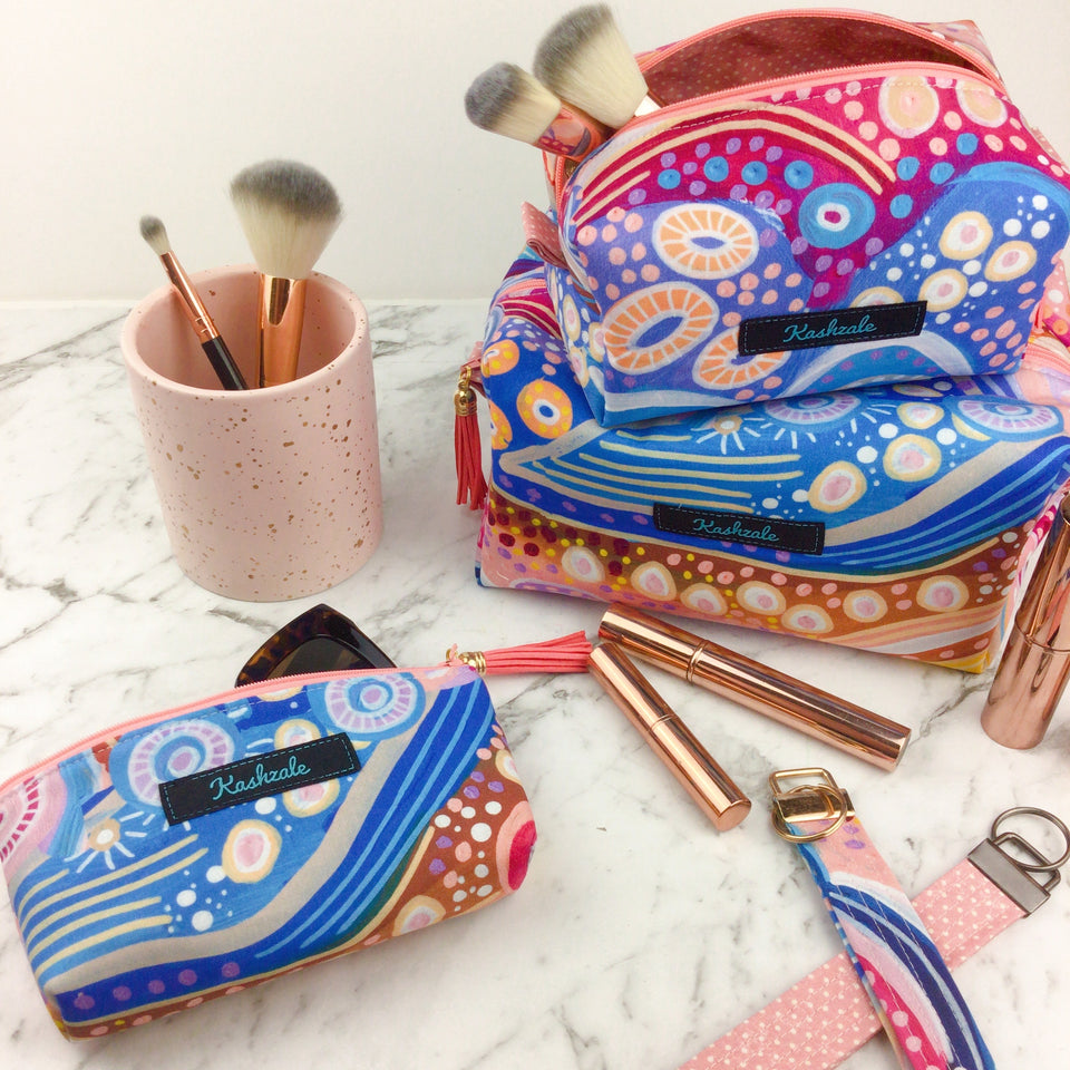 Collection of cosmetic bags featuring blue, pink and mauve Aboriginal artwork.